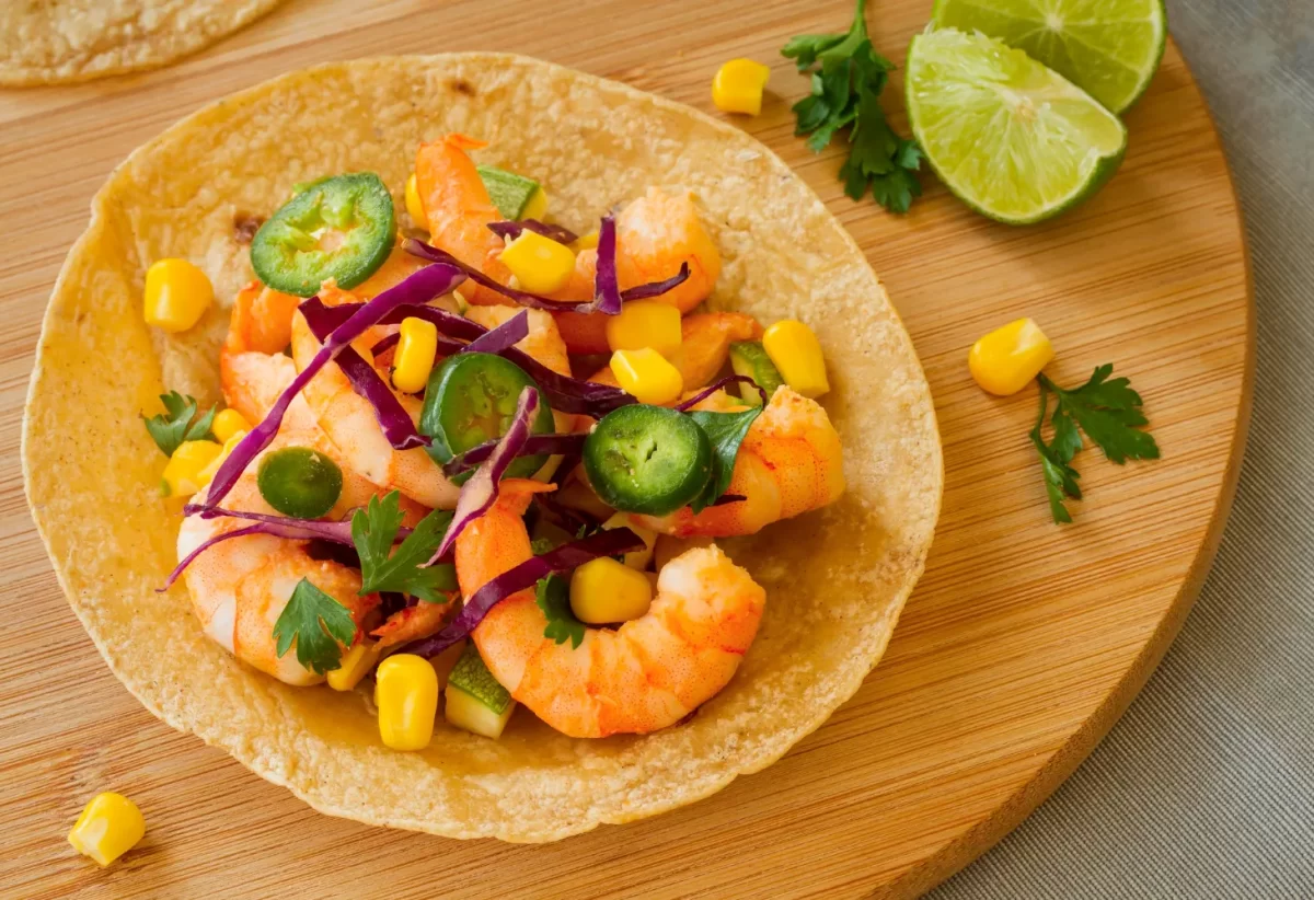 Taco de camarones con maíz, pimientos, cebolla morada y cilantro, servido en una tortilla de maíz sobre una tabla de madera. Rodajas de limón decoran el plato.