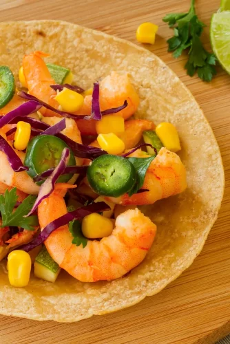 Taco de camarones con maíz, pimientos, cebolla morada y cilantro, servido en una tortilla de maíz sobre una tabla de madera. Rodajas de limón decoran el plato.