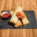 Tacos dorados servidos en una tabla de piedra, acompañados de salsa roja y decorados con una flor de cempasúchil.