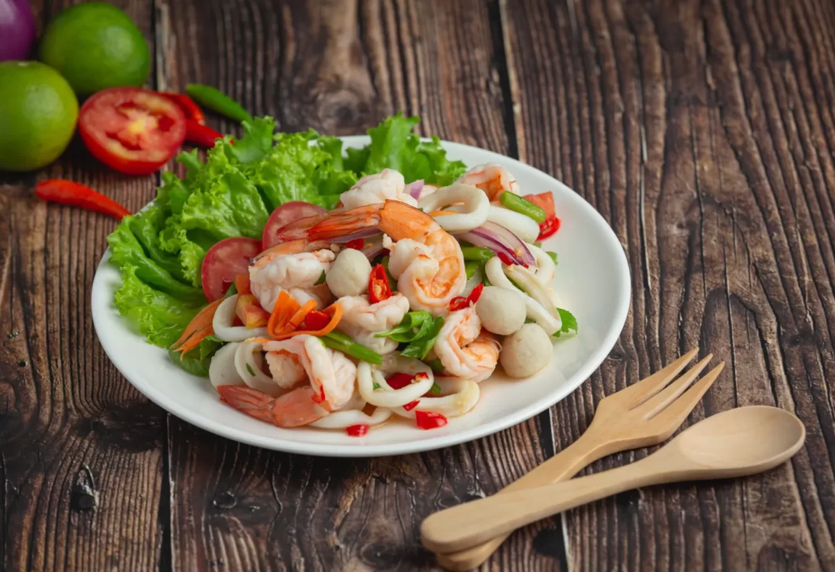 Ensalada fresca de mariscos con camarones, calamares, champiñones y verduras coloridas, servida en un plato blanco sobre una mesa de madera.