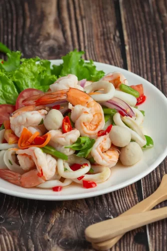 Ensalada fresca de mariscos con camarones, calamares, champiñones y verduras coloridas, servida en un plato blanco sobre una mesa de madera.