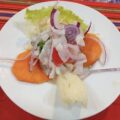 Ceviche fresco servido en un plato blanco, acompañado de lechuga, rodajas de cebolla morada y trozos de melón, con una guarnición de yuca. Ideal para disfrutar en un día caluroso.