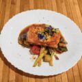 Plato gourmet de salmón glaseado servido sobre una cama de verduras asadas, incluyendo espárragos, pimientos y tomates, decorado con flores comestibles y hierbas, presentado en un plato blanco sobre una superficie de madera.