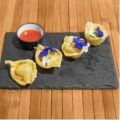 Cuatro empanaditas fritas decoradas con flores comestibles, acompañadas de una salsa roja, servidas en una tabla de pizarra sobre un fondo de madera.