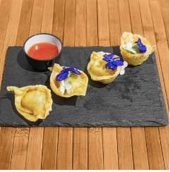Cuatro empanaditas fritas decoradas con flores comestibles, acompañadas de una salsa roja, servidas en una tabla de pizarra sobre un fondo de madera.